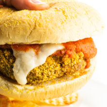 A hand holding a vegan chicken parmesan sandwich with vegan mozzarella cheese drizzling down the side and marinara sauce