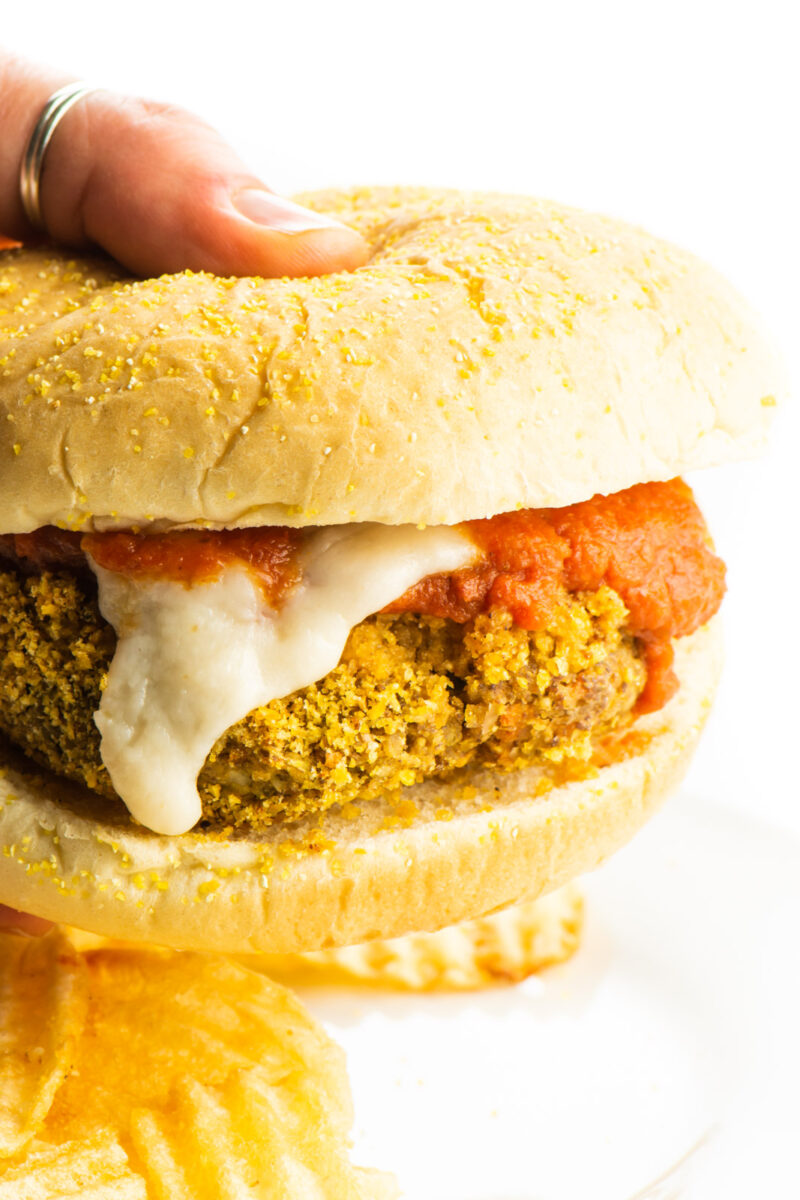 A hand holding a vegan chicken parmesan sandwich with vegan mozzarella cheese drizzling down the side and marinara sauce