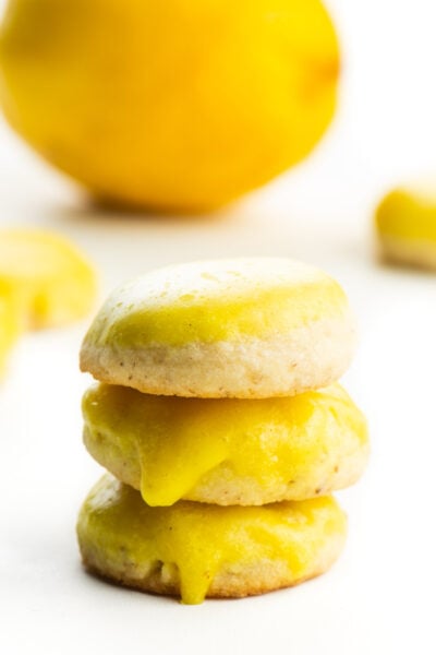 A stack of vegan lemon cookies sit in front of a lemon with other cookies around it.
