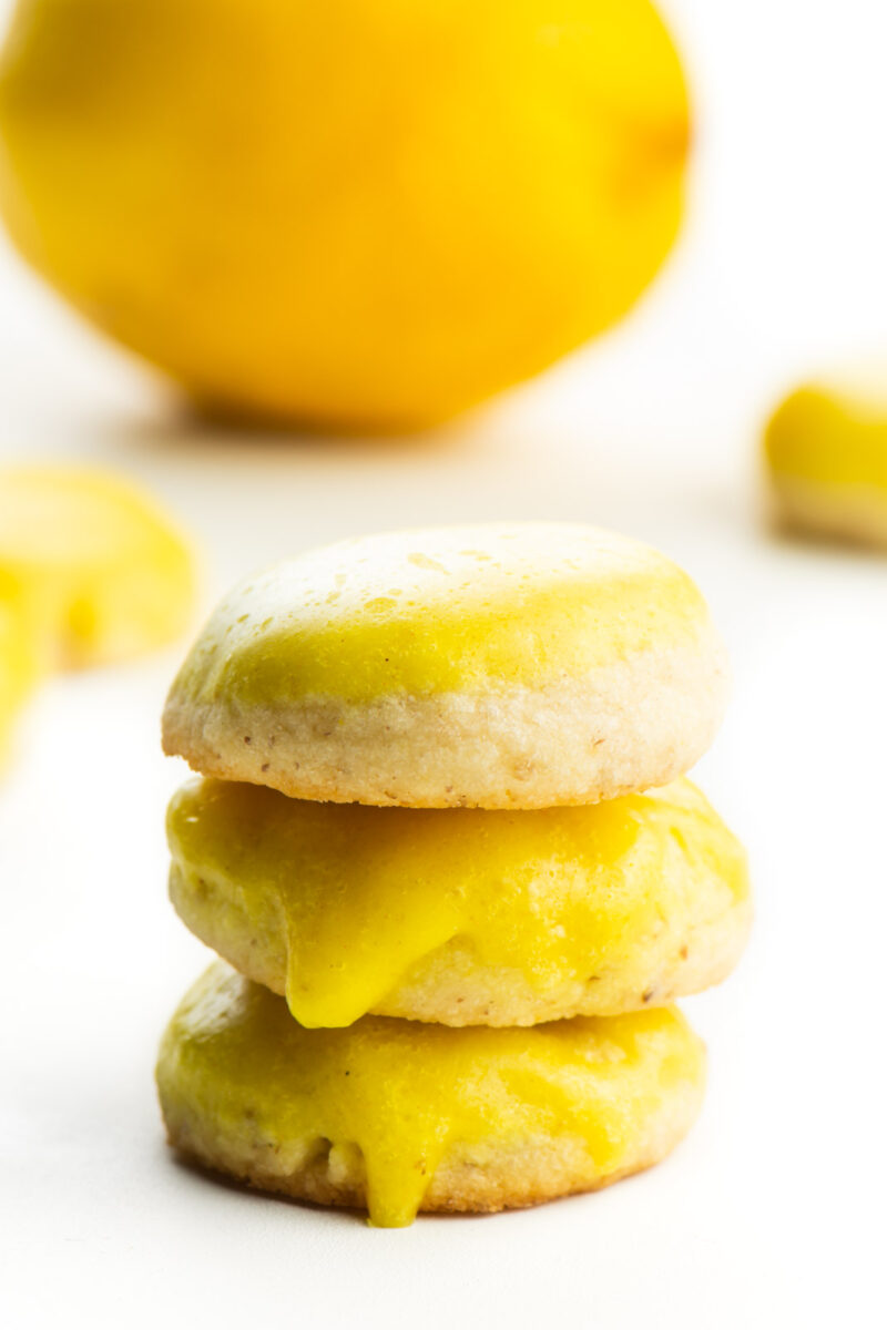 A stack of vegan lemon cookies sit in front of a lemon with other cookies around it.