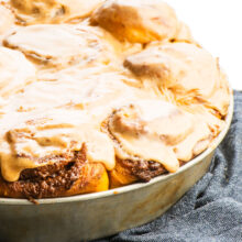 A batch of freshly baked and frosted cinnamon rolls in a pan sit on a dark blue kitchen towel.