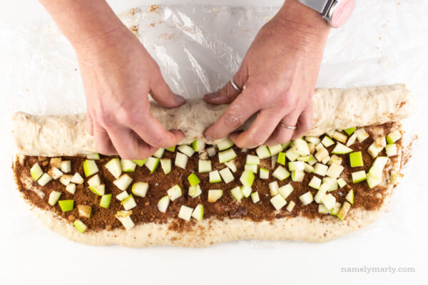 Two hands roll dough into a log to make cinnamon rolls. You can see the filling on top of the dough that has not been rolled yet.