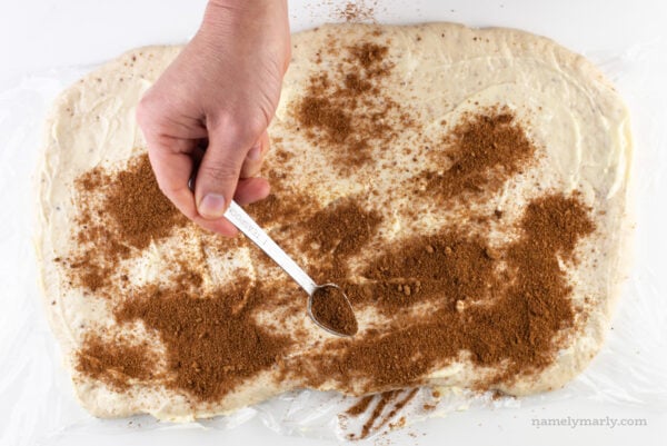A hand sprinkles cinnamon sugar over rolled out dough with vegan butter on top.