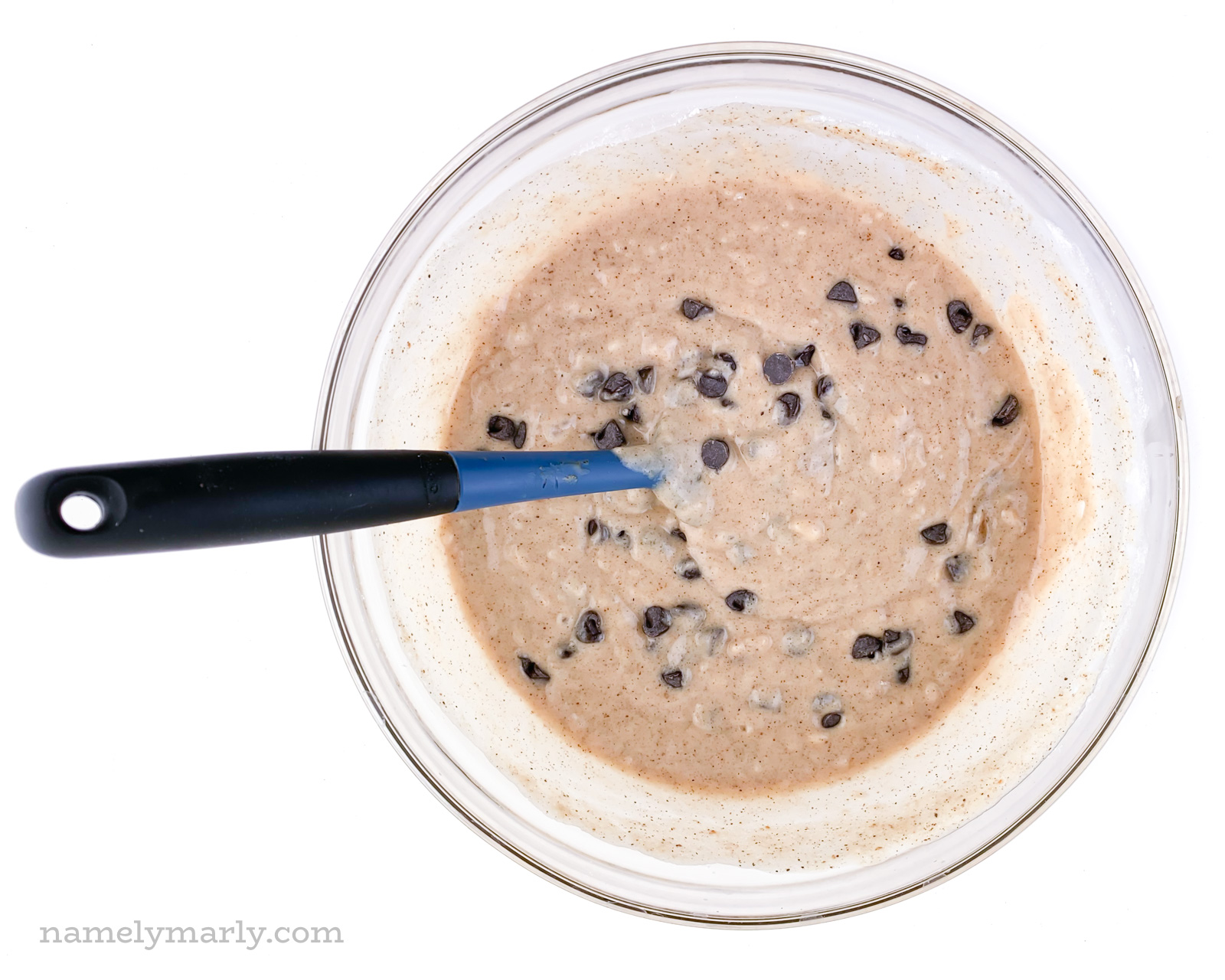 A glass bowl holds batter with chocolate chips and a blue spatula for stirring.