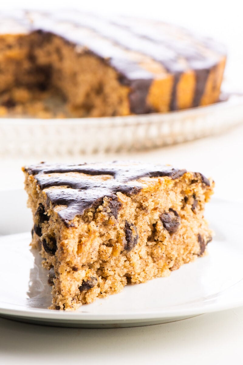 A slice of almond butter cake with chocolate drizzle on the top sits in front of the rest of the cake.