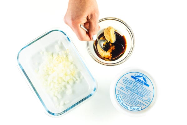 A hand is stirring marinade next to a container of miso paste and another container with chopped onions.