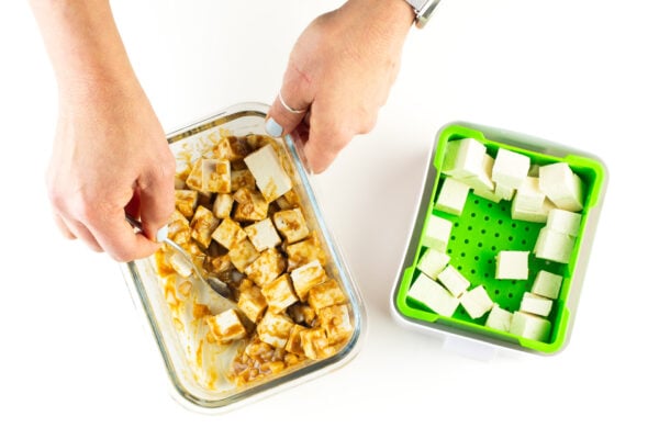A hand uses a spoon to stir tofu with marinade. The rest of the cubed tofu sits in a green container.