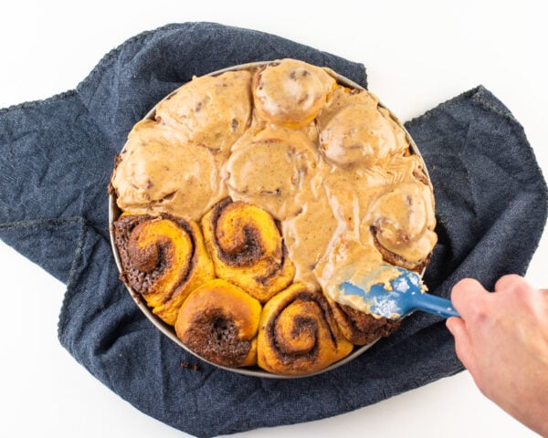 A hand holds a spatula spreading frosting over freshly baked cinnamon rolls.