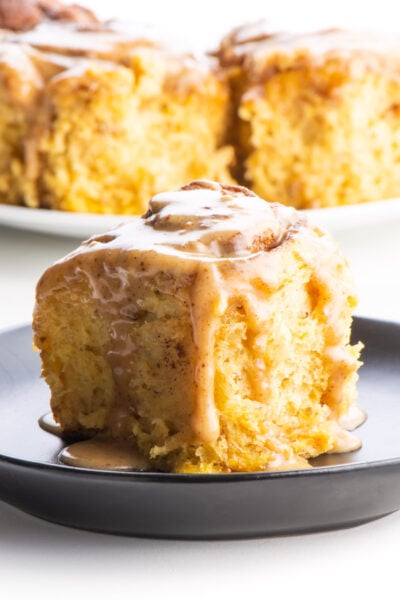 A sweet potato cinnamon roll on a black plate sits in front of a plate with several more cinnamon rolls.