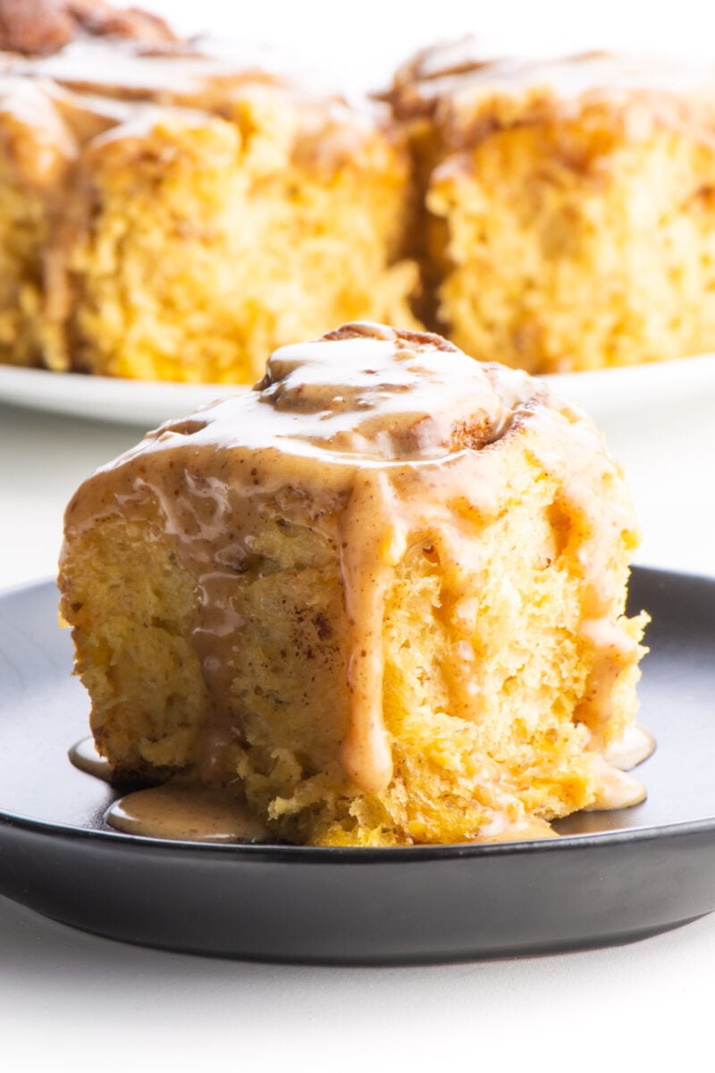 A sweet potato cinnamon roll on a black plate sits in front of a plate with several more cinnamon rolls.