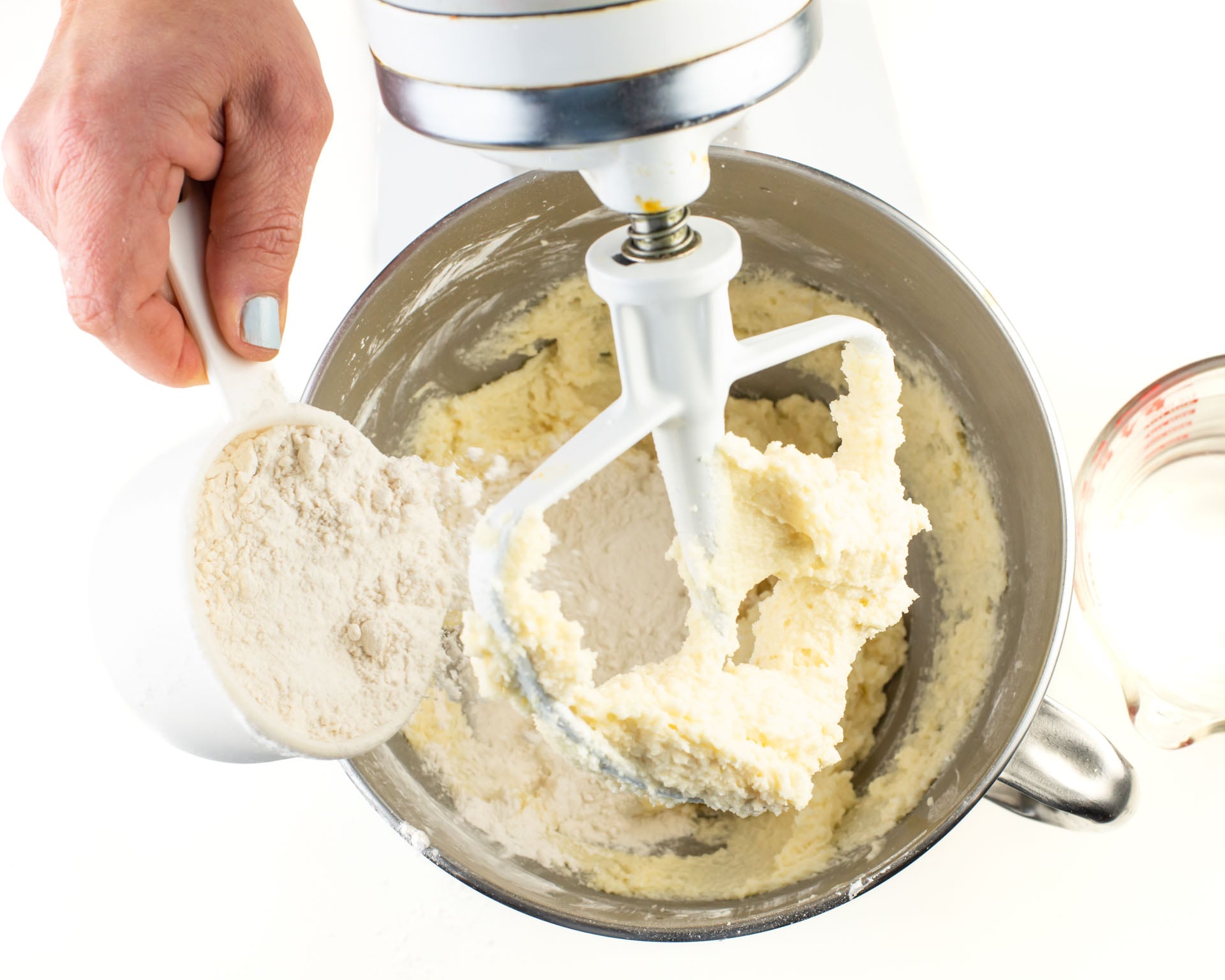 A hand pours flour into a mixing bowl. A pyrex measuring dish of water sits next to it.