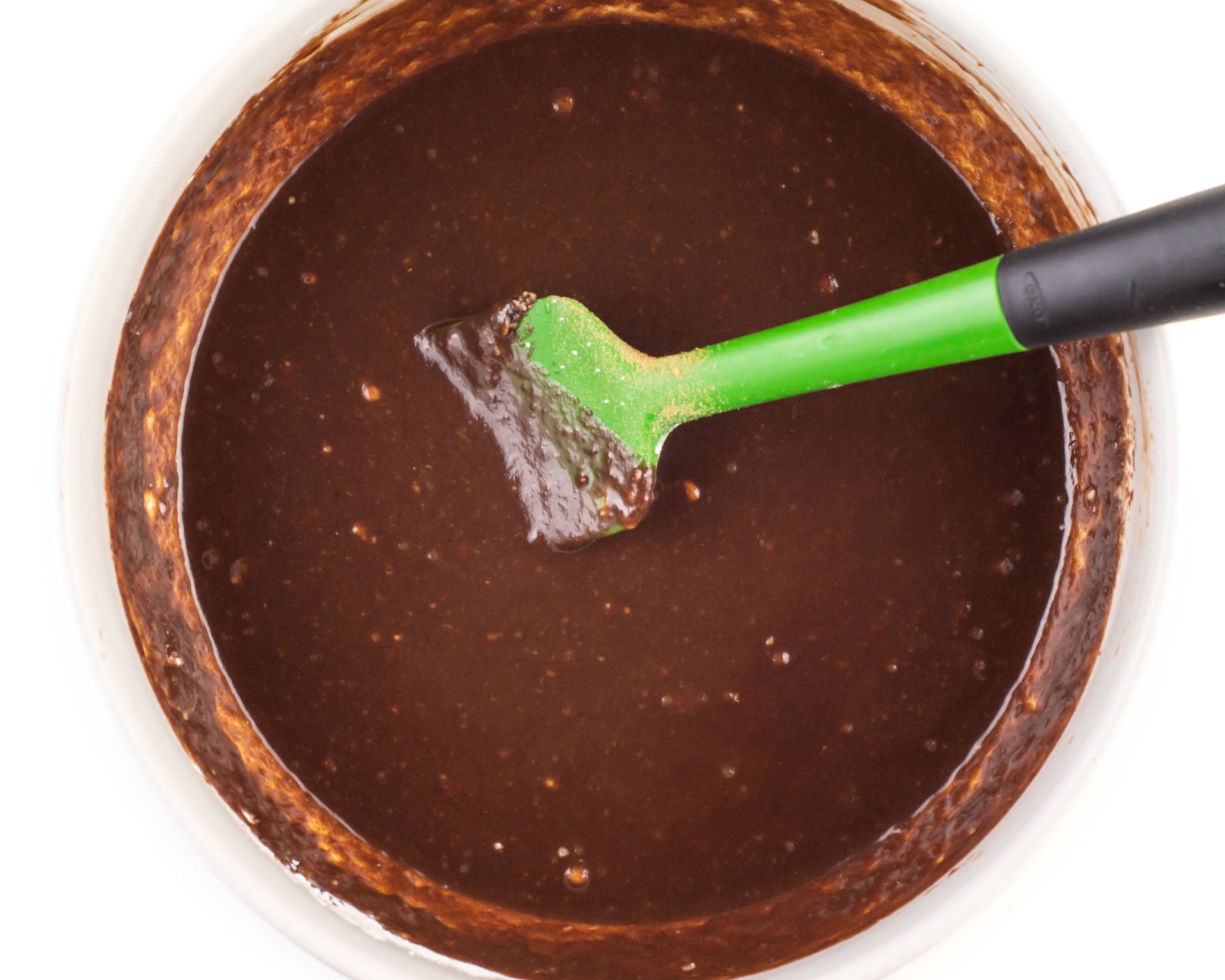 A chocolate mixture is in a glass bowl with a green spatula.