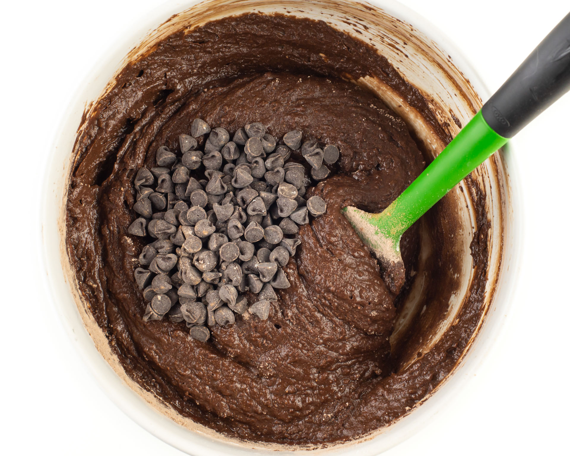 Chocolate batter is in a mixing bowl with chocolate chips. A green spatula is in the bowl.