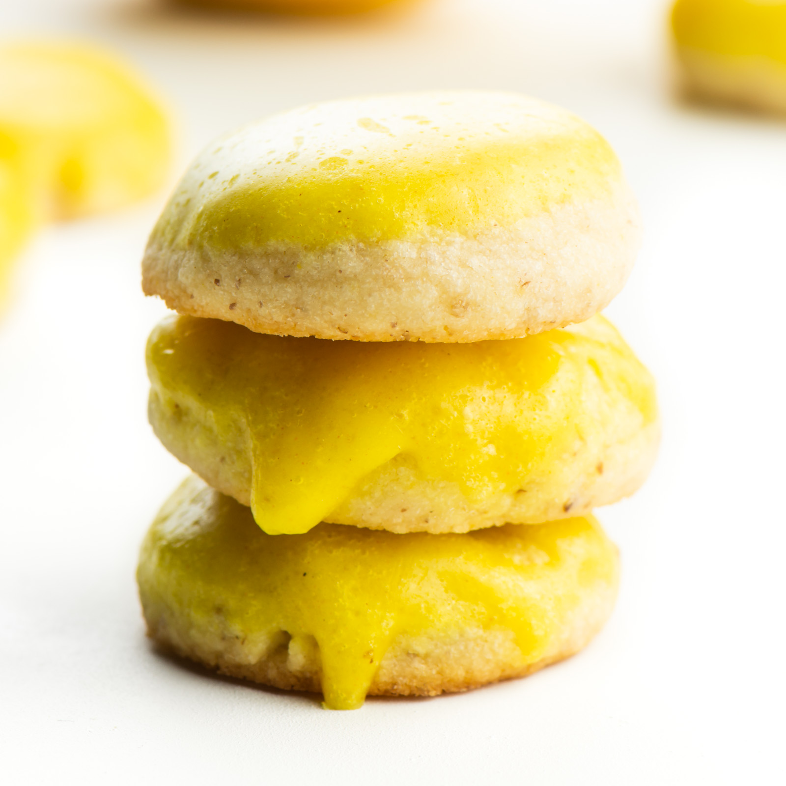 A stack of vegan lemon cookies sit in front of a lemon with other cookies around it.