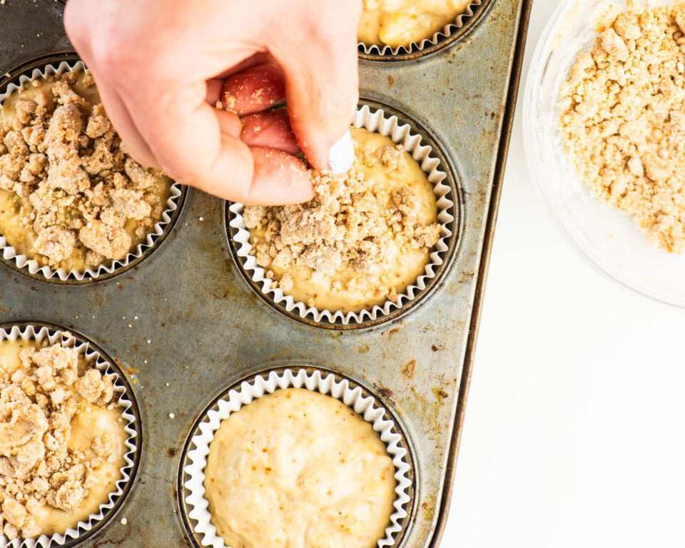 A hand is placing streusel crumbles on top of muffin batter in a muffin tin.