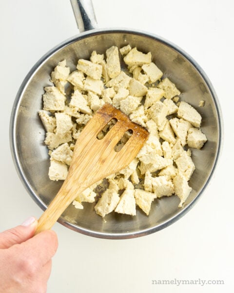 Tofu is in a skillet and a hand holds a spatula breaking the tofu into smaller pieces.