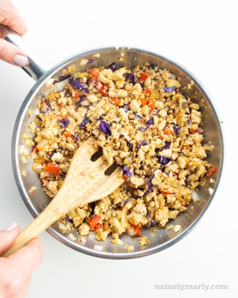 Tofu filling with veggies is in a skillet and a hand holds a spatula stirring it.