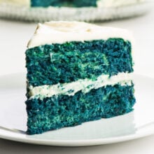 A slice of blue velvet cake on a plate in front of the rest of the cake.