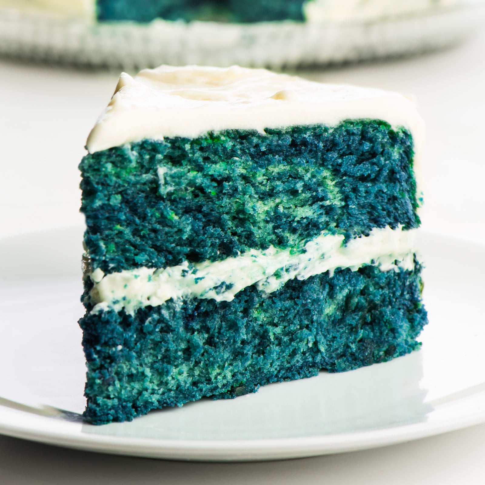A slice of blue velvet cake on a plate in front of the rest of the cake.