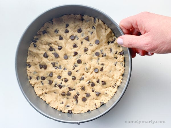 A hand holds a pan full of cookie cake batter with chocolate chips pressed into the top.