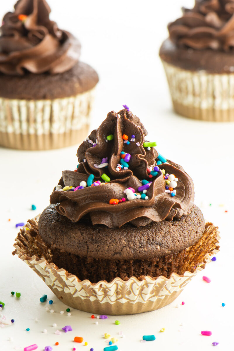 A cupcake topped with vegan chocolate frosting and sprinkles sits in front of 2 other cupcakes.