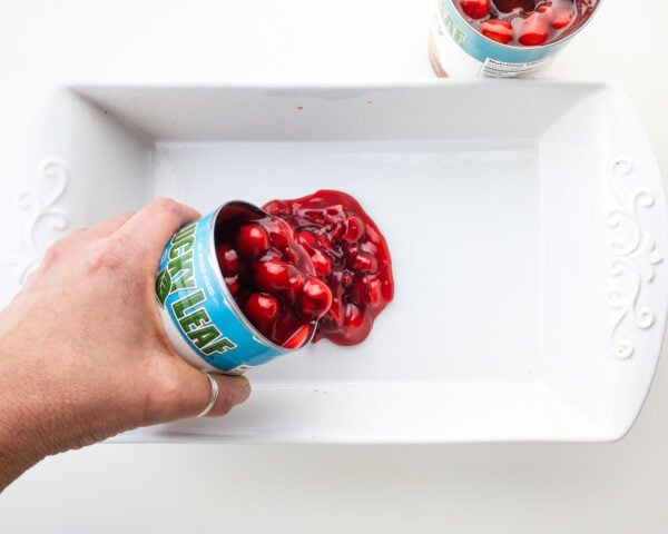 A hand pours a can of cherry pie filling into a white baking dish.
