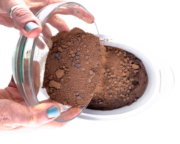Two hands hold a glass bowl pouring chocolate crumbles over a cake batter.