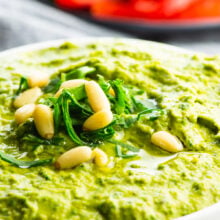 A closeup of green hummus topped with chopped greens and pine nuts and a plate of sliced red bell peppers behind it.