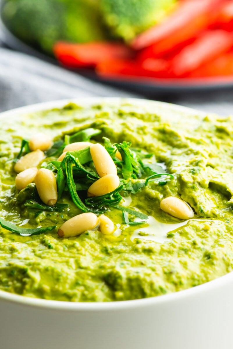 A closeup of green hummus topped with chopped greens and pine nuts and a plate of sliced red bell peppers behind it.