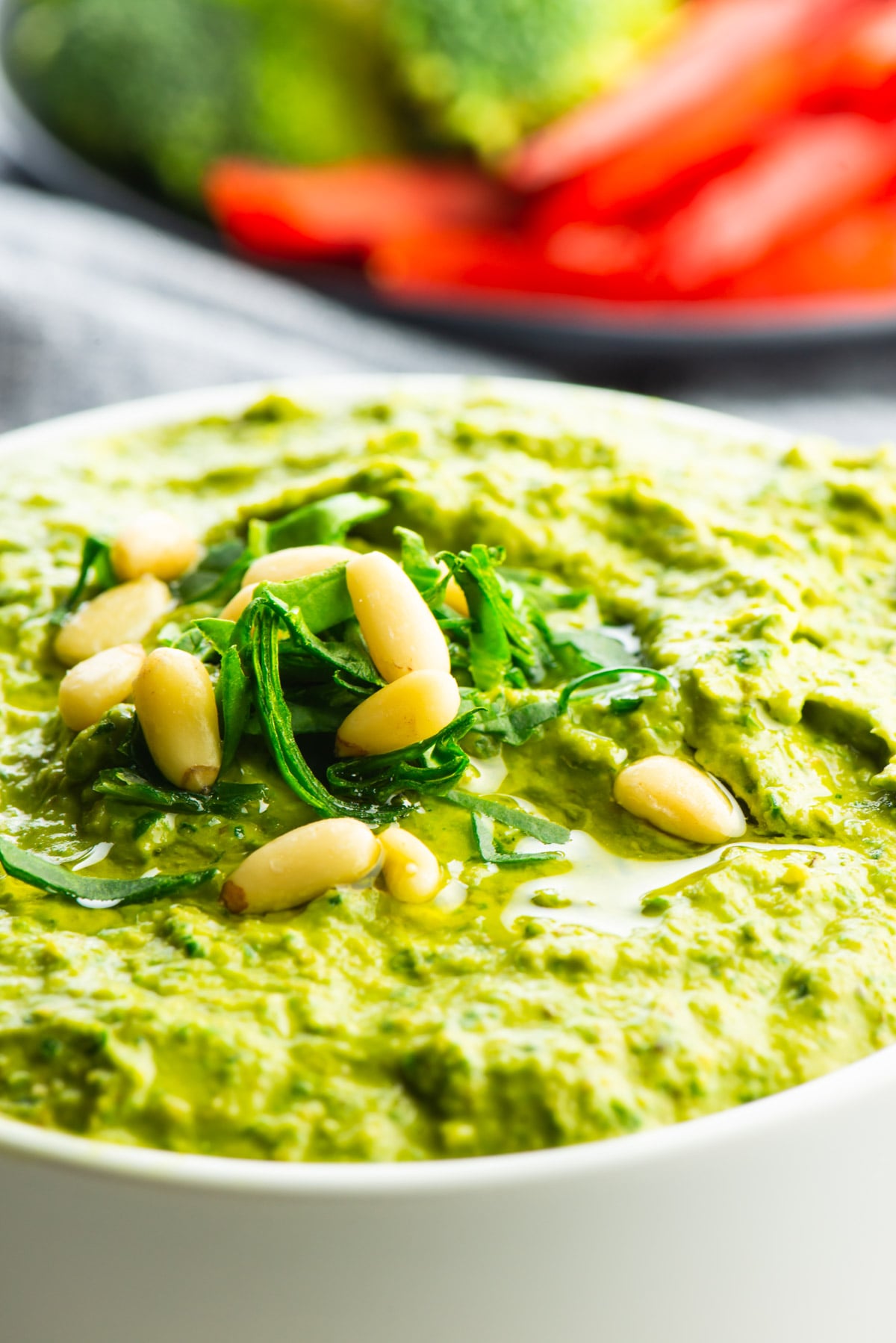 A closeup of green hummus topped with chopped greens and pine nuts and a plate of sliced red bell peppers behind it.
