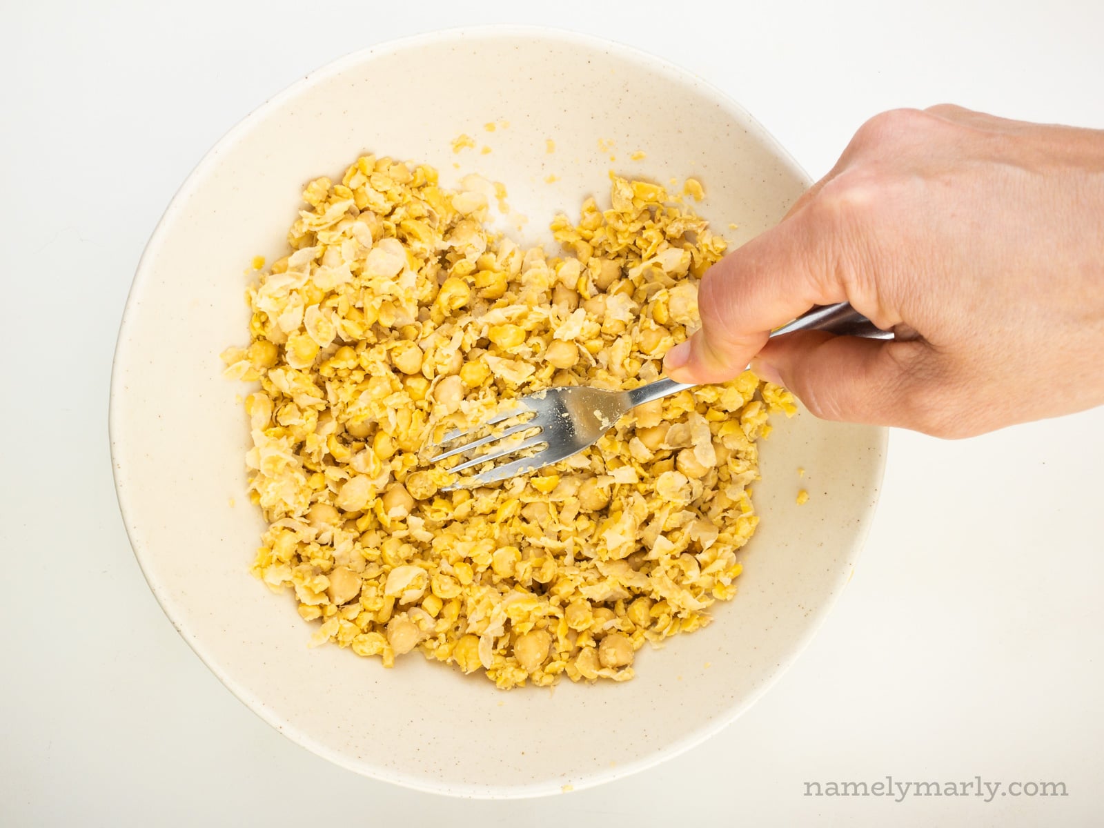 A hand holds a fork mashing chickpeas in a bowl.