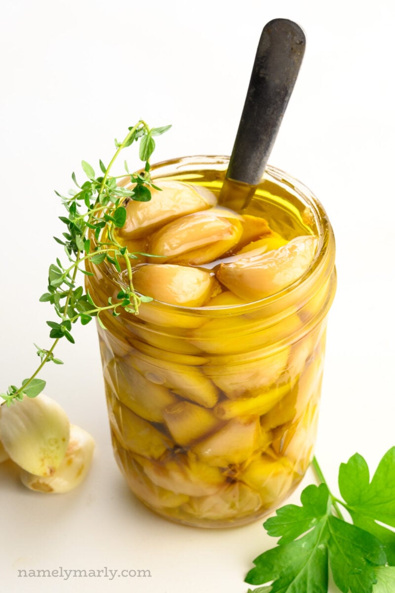 A mason jar holds garlic confit with a spoon. There are green herbs on top of and beside the jar.