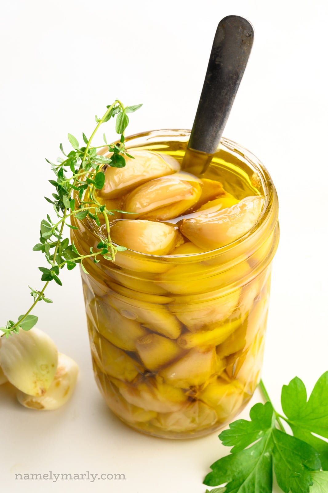 A mason jar holds garlic confit with a spoon. There are green herbs on top of and beside the jar.