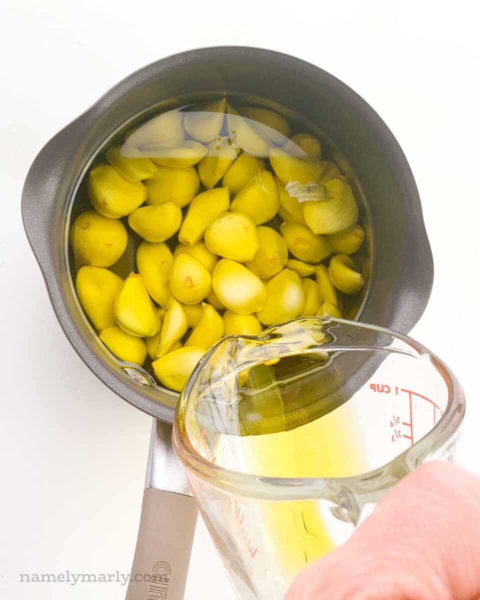 A glass measuring cup is pouring olive oil in a saucepan full of garlic cloves.