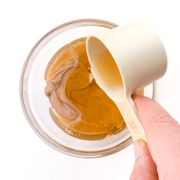 A hand holds a measuring cup full of syrup and is pouring it over a glass bowl full of peanut butter.