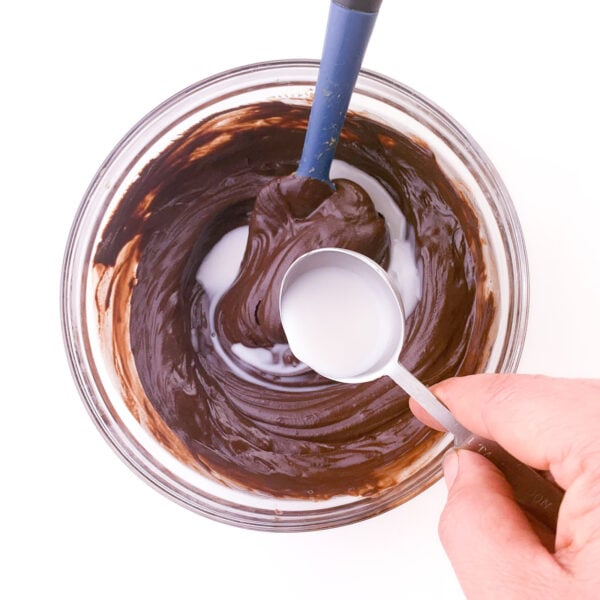 A hand holds a spoon pouring milk into a bowl with melted chocolate. There is a blue spatula in the bowl too.