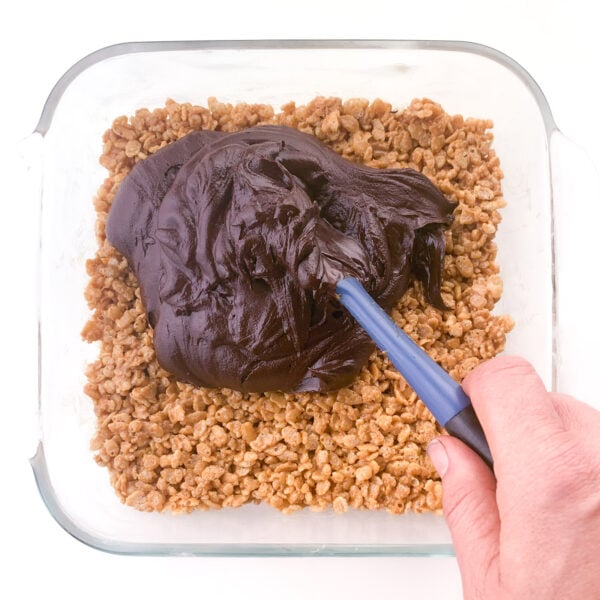 A hand holds a spatula and is spreading soft chocolate ganache over peanut butter rice crispies bars.