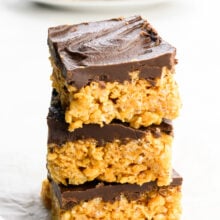 A stack of three peanut butter rice krispies treats topped with chocolate ganache and more treats on a plate behind it.
