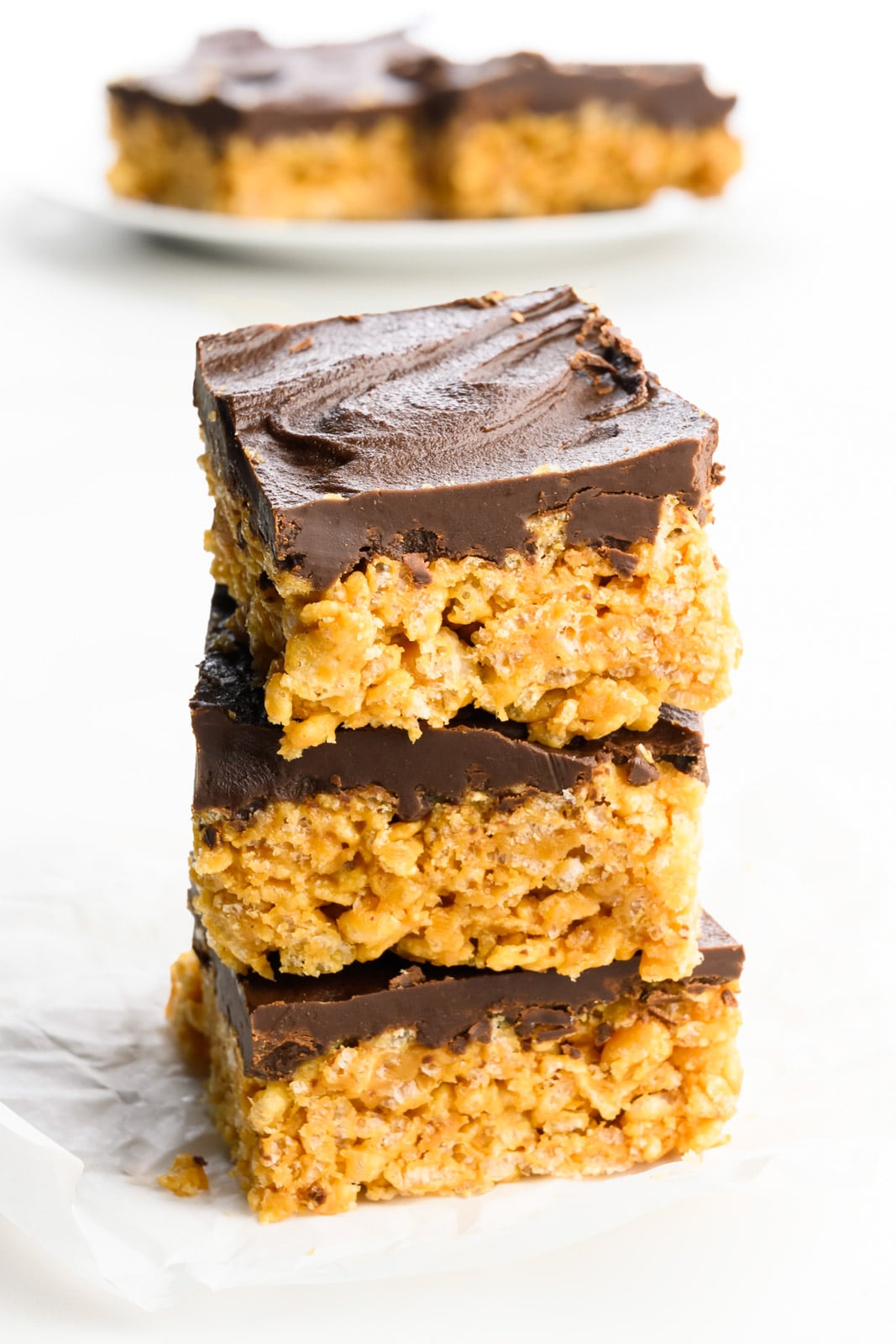 A stack of three peanut butter rice krispies treats topped with chocolate ganache and more treats on a plate behind it.