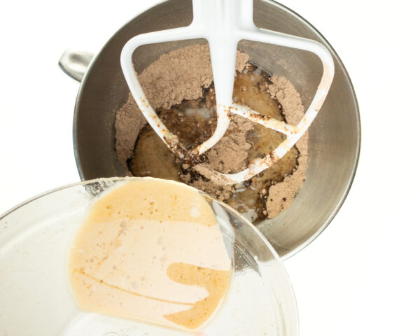 Wet ingredients are in a bowl and poured into a mixing bowl of a stand mixer with dry flour ingredients.