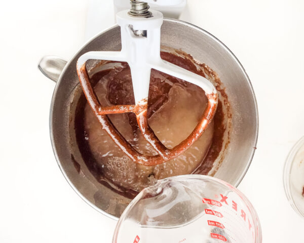 Hot water in a pyrex measuring cup is being poured into a mixing bowl of a stand mixer with chocolate cake batter in it.