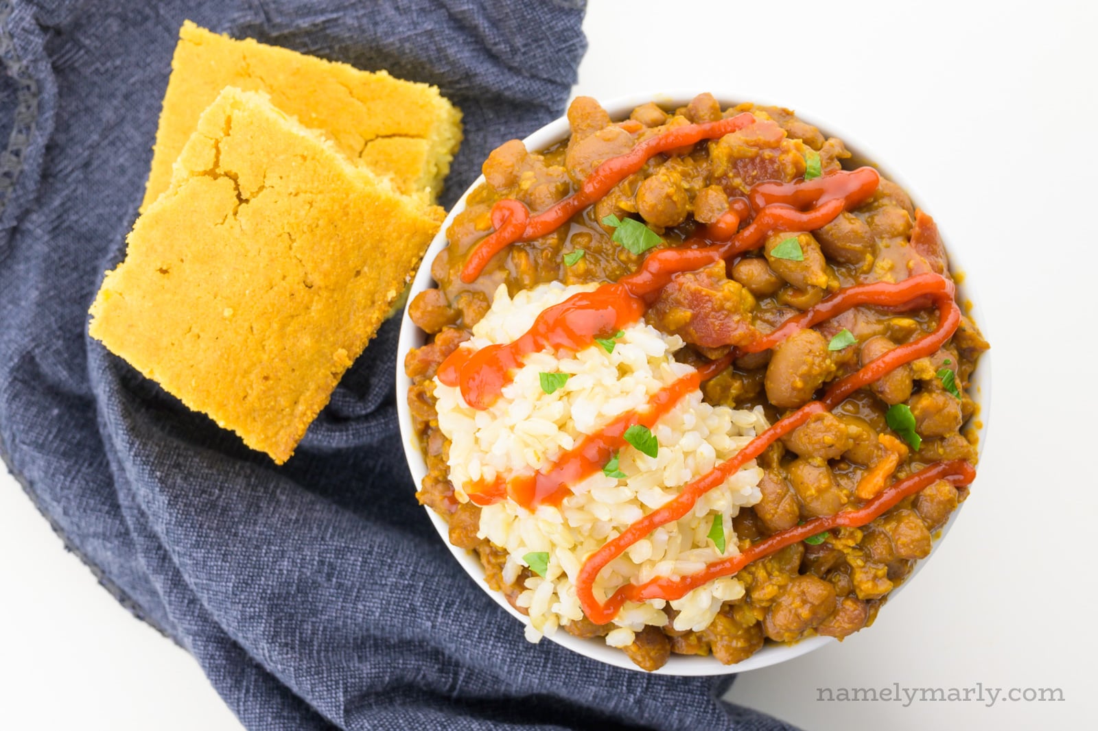 Looking down on a bowl of beans and rice with cornbread beside it. Hot sauce has been drizzled over the top.