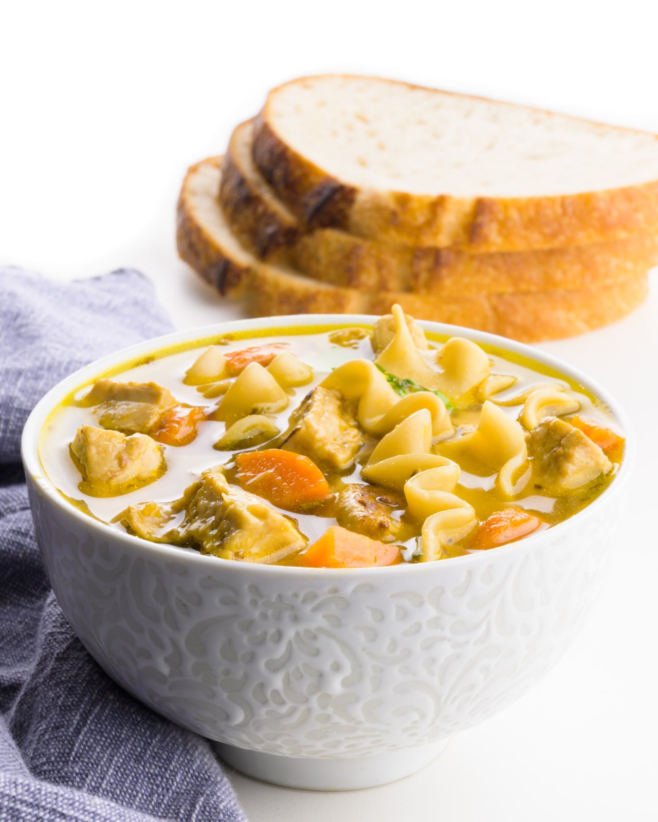 A bowl of soup has a blue napkin beside it and sliced bread in the background.