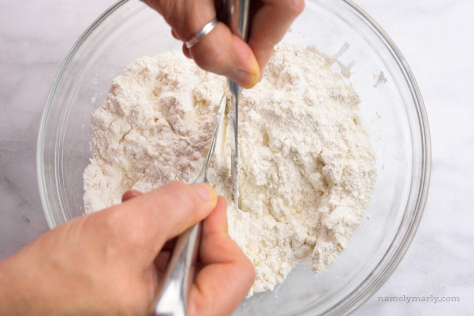 Two hands are holding butter knives and cutting in butter in flour.