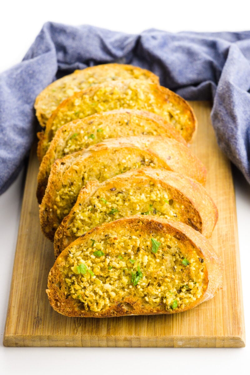 Several slices of vegan garlic bard are on a cutting board with a blue kitchen towel behind it.