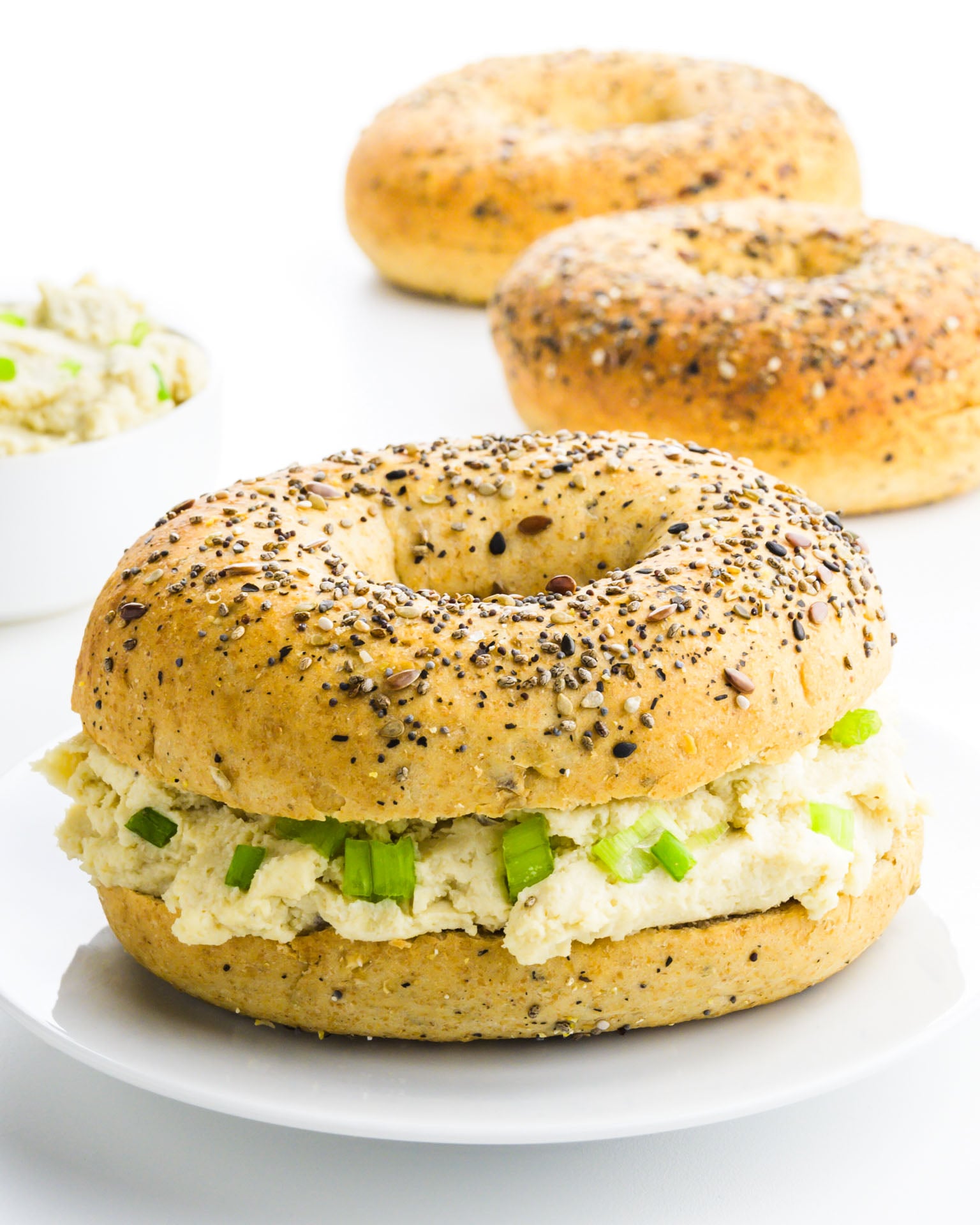 A bagel has been cut in half and has cashew cream cheese in the middle. There are more bagels behind it and a bowl of more vegan cream cheese.