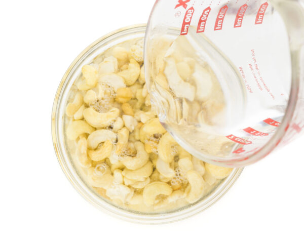 A bowl of raw cashews has been filled mostly with water. A pyrex measuring cup is finishing up pouring water over it.