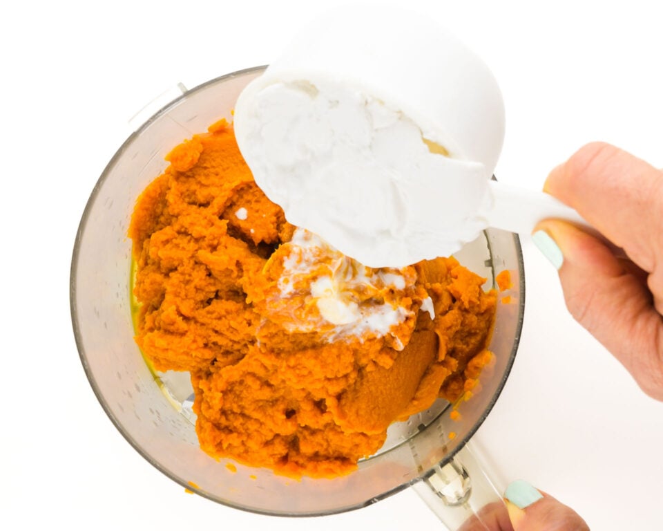 Coconut milk is being poured into a food processor bowl full of pumpkin puree.