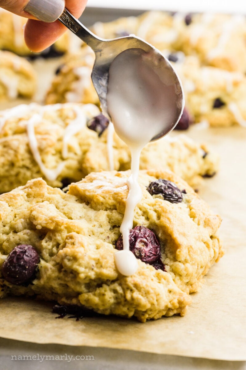 A hand holds a spoon and drizzles frosting over scones.