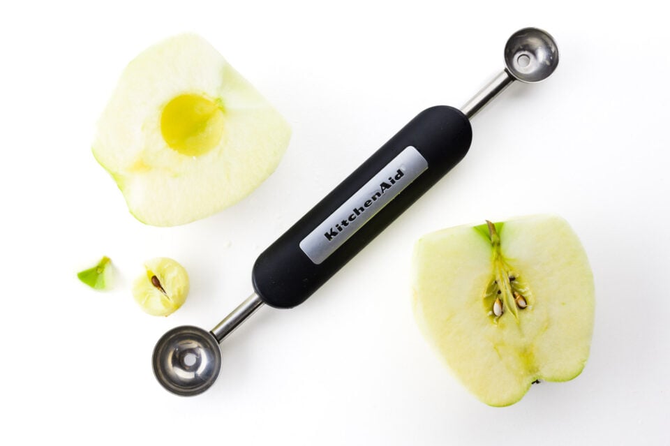 A melon baller sits between to halves of an apple. One has a core and stem bits removed.